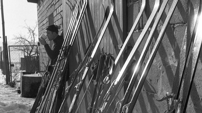 A man leans to a shed drinking a cup of coffee next to several skis also leaning to the shed.