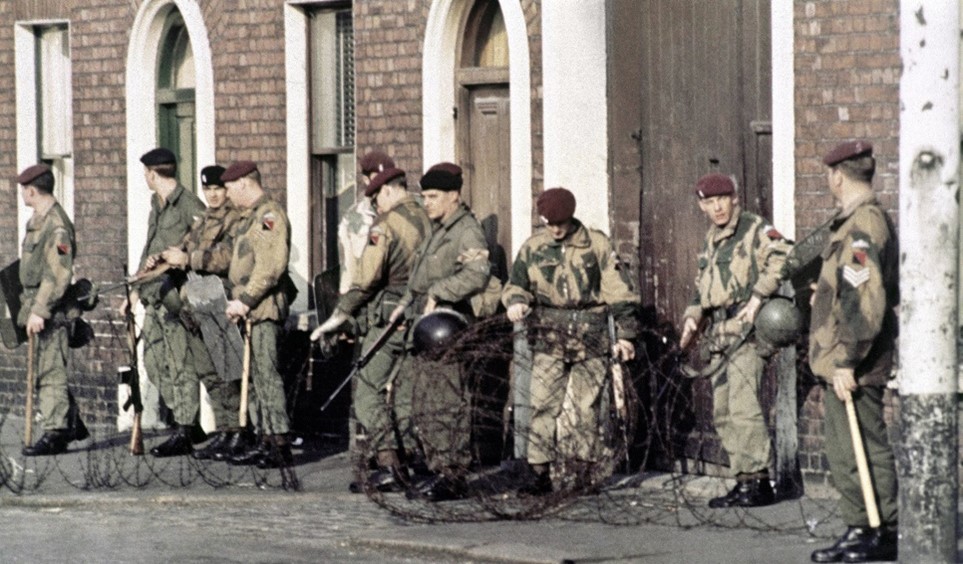 A color photograph of a street in Northern Ireland.  Brick buildings with arched doorways are in the background.  A line of soldiers in camouflage uniforms and red berets stand facing toward the left behind a stretched-out roll of barbed wire.  The soldiers are carrying batons and some are carrying heavier helmets.