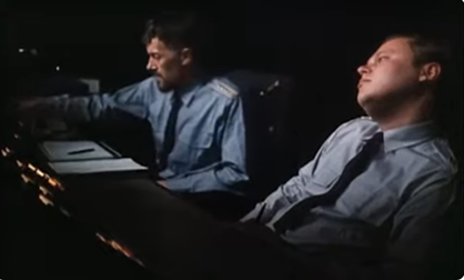 A color still from the movie The Peace Game of two white men , the controllers, in blue button down shirts and black ties.  The man on the left is sitting up and reading a document on a desk in front of him and reaching for a button on the control panel, while the man on the right slouches back in his chair, staring blankly.