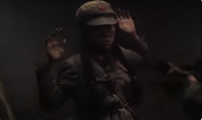 A dimly-lit color still from the movie The Peace Game. A woman in a Red Army uniform and cap faces slightly to the right of the viewer, her hands held up in surrender.