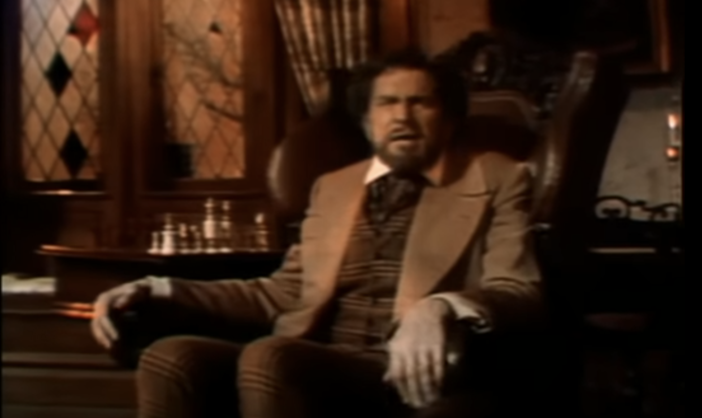 A film still of Vincent Price, this time with dark fluffy hair and a van dyke beard. He is wearing a brown sport jacket over a brown plaid waistcoat with matching brown plaid trousers and necktie. He is sitting in a brown leather wingback armchair with his hands gripping the ends of the chair arms, looking at the viewer.  The chair sits in front of a round side table with glassware on it.  In the background is a pair of diamond lattice windows, of which three diamonds have colored glass instead of clear.