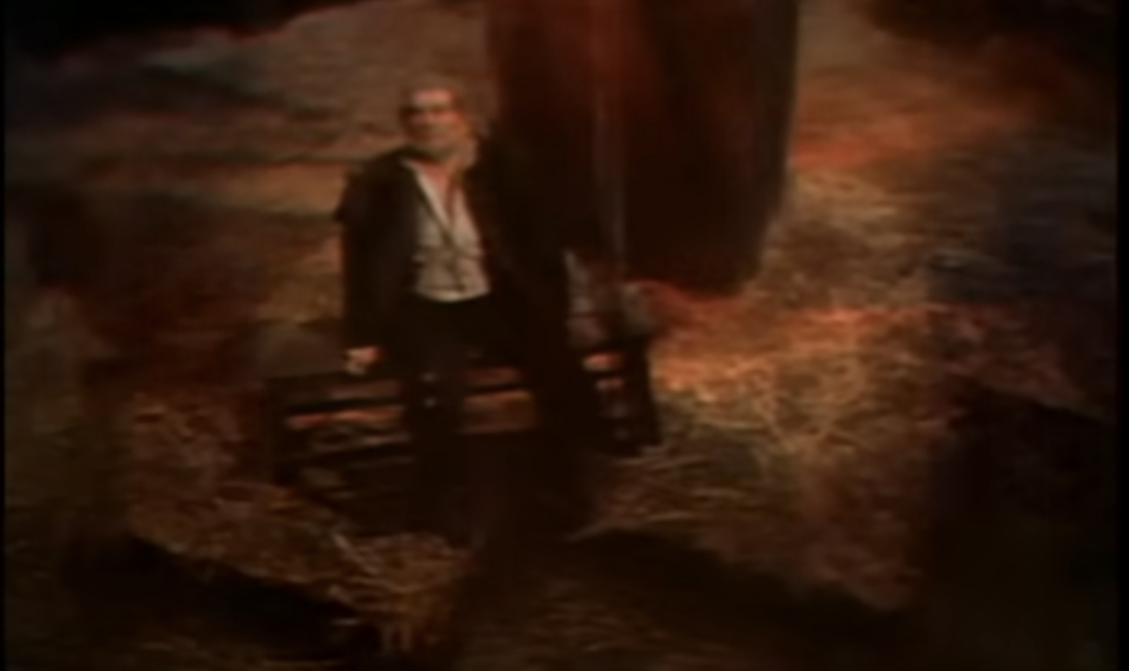 A blurry film still of Vincent Price sitting on a bench at the bottom of the pit, looking up at the viewer.  There is a large support column behind him and straw covering the floor. A blurry line that may be the pendulum is to the right of the frame, as though it had just swung past Price's face.