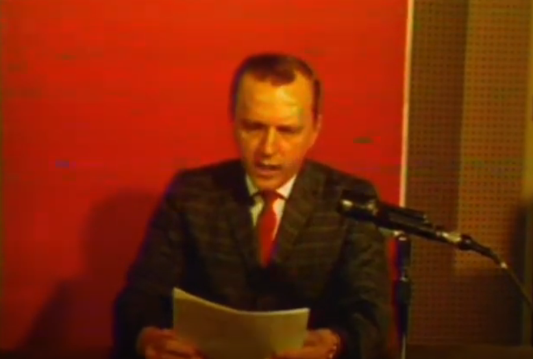 A color film still of a white  man with brown hair wearing a plaid suit and red tie sitting in front of a red background with brown acoustic tile on the right side.  He is holding a stack of papers from which he is reading into a black microphone.