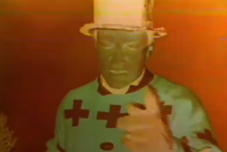 A negative image of a color still.  A man in a top hat wearing a cardigan is standing in front of an orange background gesticulating at the screen.  The hat is white instead of black, the man's face green, and the cardigan a bright teal with black +s.