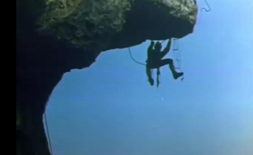 A color still of a person hanging from the underside of a cliff overhang. The person is supported by several ropes and a rope ladder.