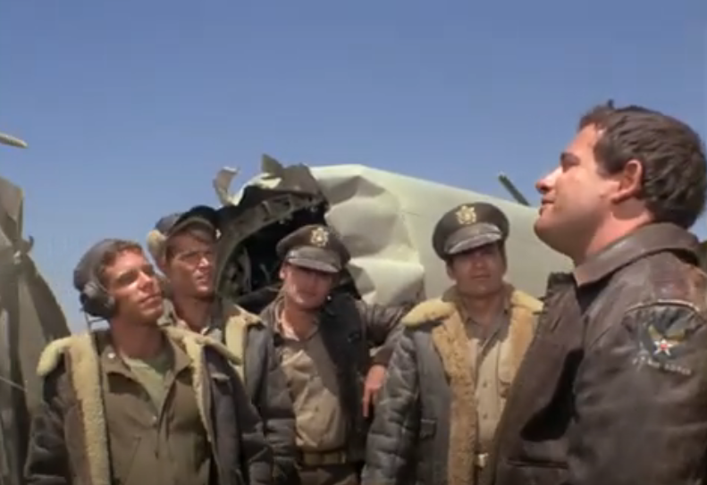 A color film still of five white men in World War II era military bomber jackets. They are standing in front of pieces of wrecked airplane.  The man on the left wears a flight helmet. The man next to him wears a baseball cap.  The third and fourth men are wearing officers' caps. All four look at the fifth man on the far right of the frame, who is facing them and appears to be speaking.