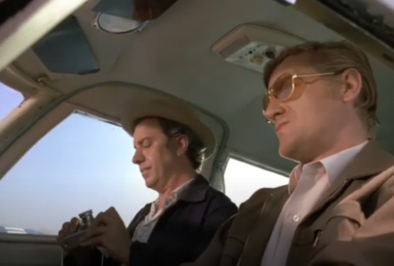 Two white men sitting in the tan interior of a small plane.  The passenger wears a tan hat, a white shirt, and dark jacket, and is adjusting a camera. The driver wears sunglasses, a pink shirt, and a brown jacket.