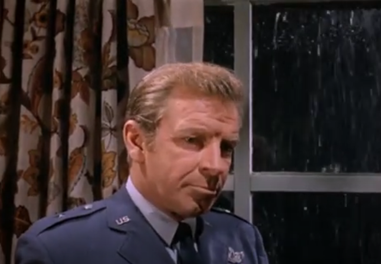 A blonde white man in general's dress uniform stands in front of window with a flower-print curtain.  The man looks pensive.  The view outside the window is dark and rainy.