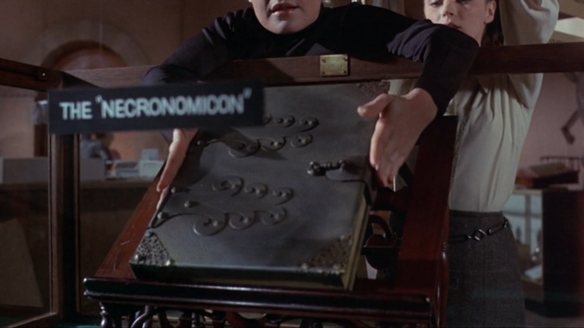 A color film still. A man in a black shirt, his face visible only from the mouth down because of the camera angle.  He reaches into a glass case to gently lift out a large black hardbound book with metal fittings and a lock on the pages. The label at the top of the case reads 'The 'Necronomicon' ' in white capitals. A woman in a white shirt stands behind the man, her hands raised, watching what he is doing.  Her face is visible only from the eyes down.