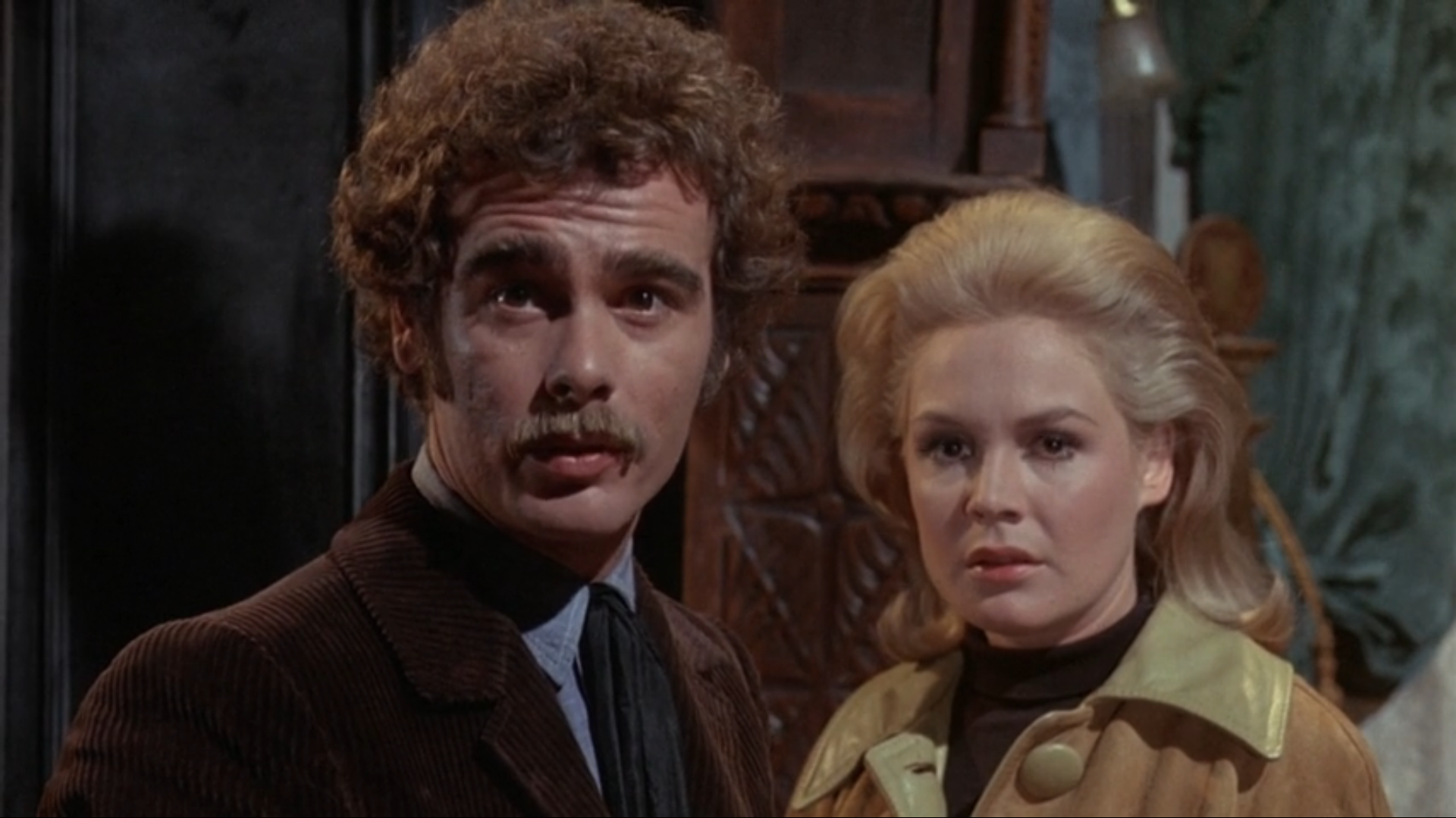 A color film still of a young white man with brown curly hair and a mustache and a young white woman with blonde hair.  They are staring to the left of the viewer with apprehensive looks on their faces.  The man wears a blue shirt, dark tie, and brown jacket.  The woman wears a black blouse and tan jacket.  Some dark wooden furniture is behind them, along with teal wallpaper.