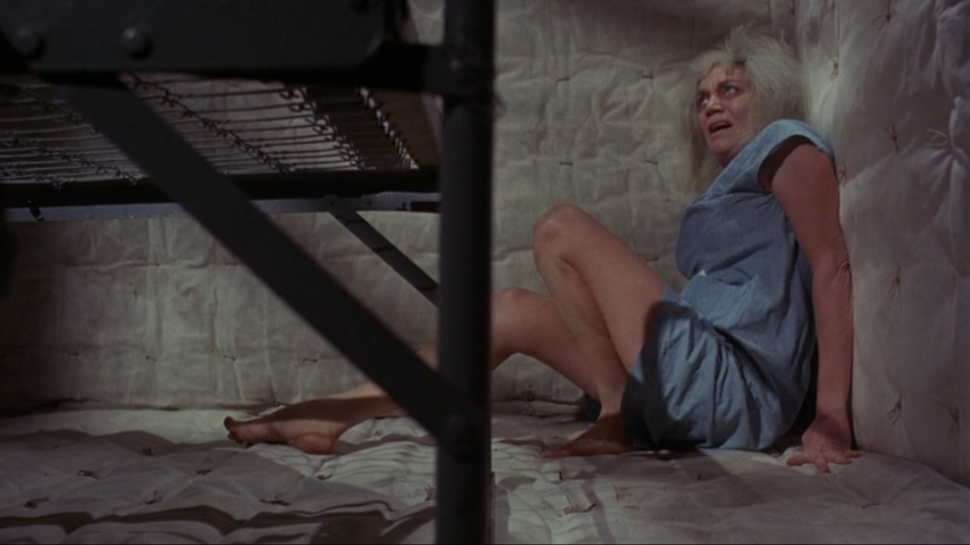 An old woman with unkempt white hair and a blue one-piece hospital gown huddles in the corner of a padded cell looking upward with a frightened expression.  The buttons of the padding - like on a mattress - are visible on the walls and floor. The metal frame and springs of a bed are in the foreground.