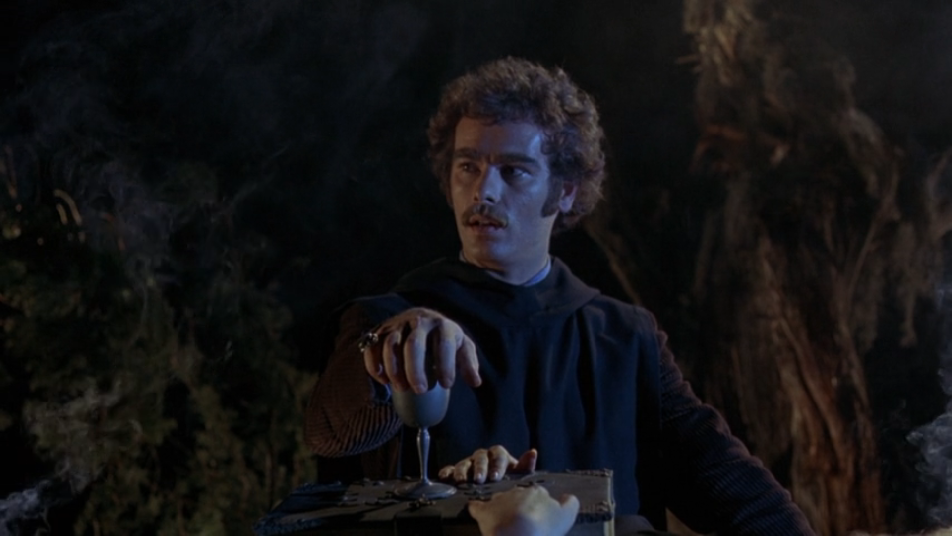 A white man with brown curly hair - the same one from the earlier photo - stands among trees at night.  He looks off to the left, seemingly in concentration.  He is  wearing a black cultist robe and rests his hand on top of a metal wine goblet which is standing on a wooden board.  A hand, presumably attached to a body lying on the board, is visible at the bottom of the screen.