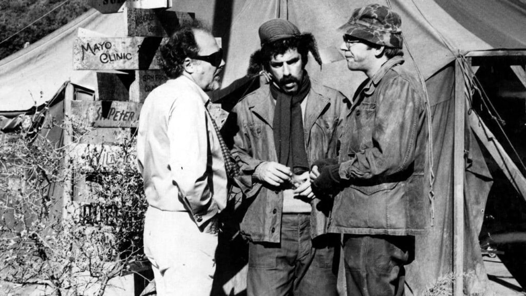 a small wide man in sunglasses is speaking to two taller actors in army fatigues