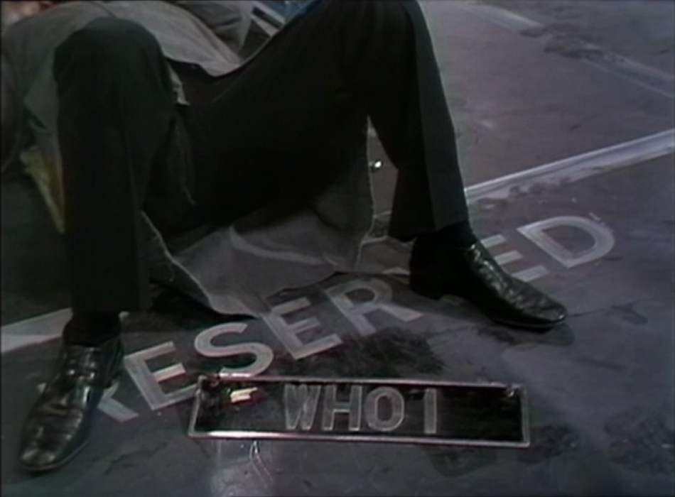 The Doctor's lower body sticking out from under his car (offscreen) as he works on it. There is a number plate at his feet with 'WHO 1' as the registration.