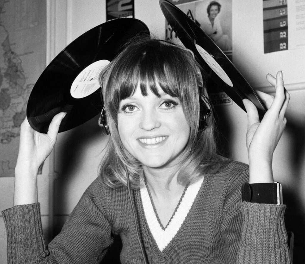 Annie Nightingale in 1970 wearing headphones and holding two records above her head in each hand.