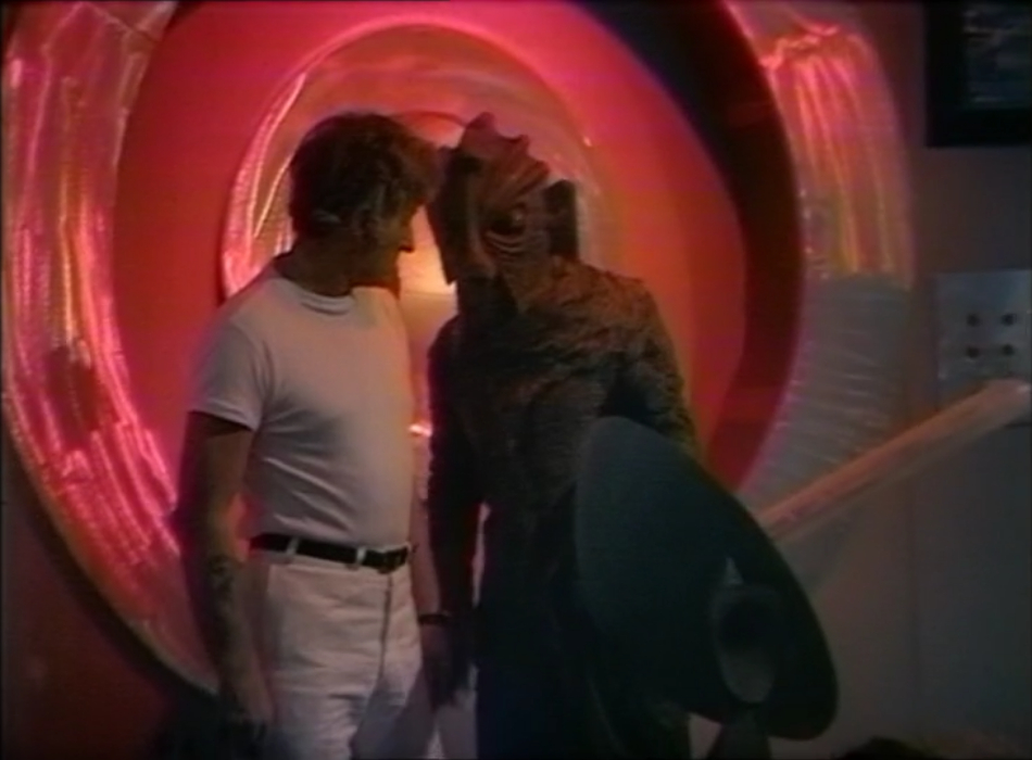 The Doctor and the Younger Silurian standing in front of the cyclotron. It's glowing, red, and circular. The Doctor has taken off his lab coat and is wearing a white t-shirt and trousers.