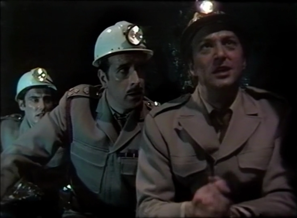 The Brigadier kneels in a dark cave with a pair of UNIT soldiers. All three look worried and are wearing tan military overalls and white hard hats with lamps attached.