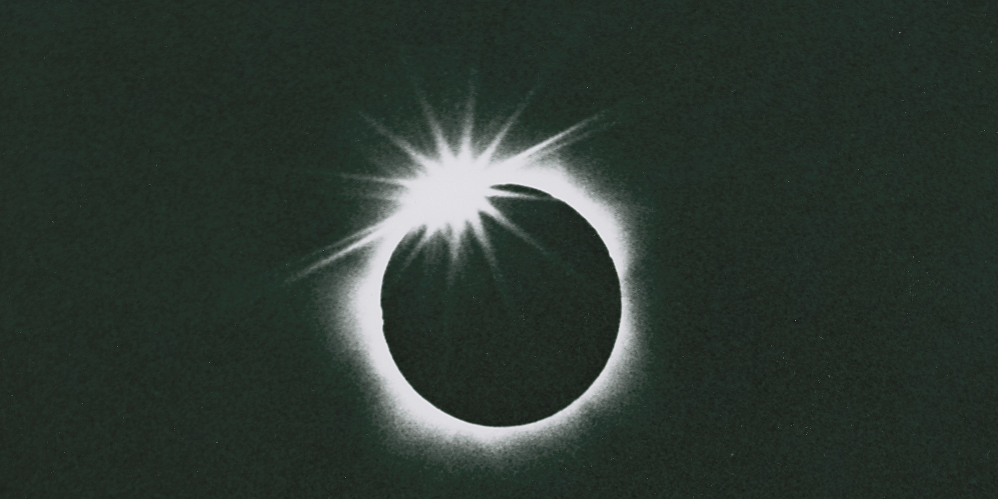 Black and white photograph with the silhouette of the moon centered and the haze of the corona seething around it. The final sliver of sunlight gleams like a gem at the top left of the 'ring'