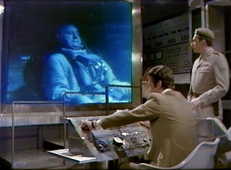 Cornish and the Brigadier watch from mission control as the Doctor appears on a big blue screen. His face is contorted by extreme g-force.