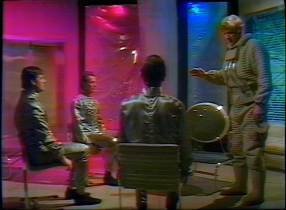 The Doctor talks to the three astronauts in a room lit in pink and blue. There's a space-age television set with a blank screen and the astronauts are all seated and wearing silver suits.