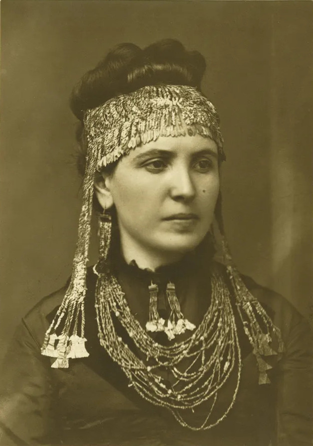 Sepia-toned portrait of a dark-haired white woman wearing many strands of beads, a beaded and plated cowl, and long narrow beaded pendant earrings