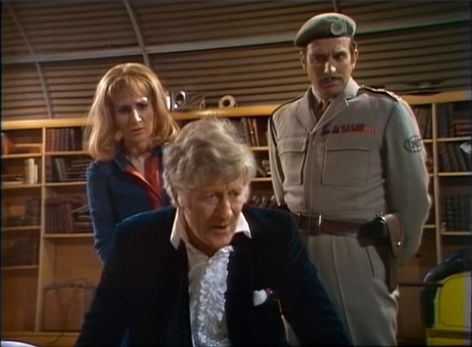 Liz and the Brigadier regard the Doctor with concerned looks on their faces as he sits with his back to them. 