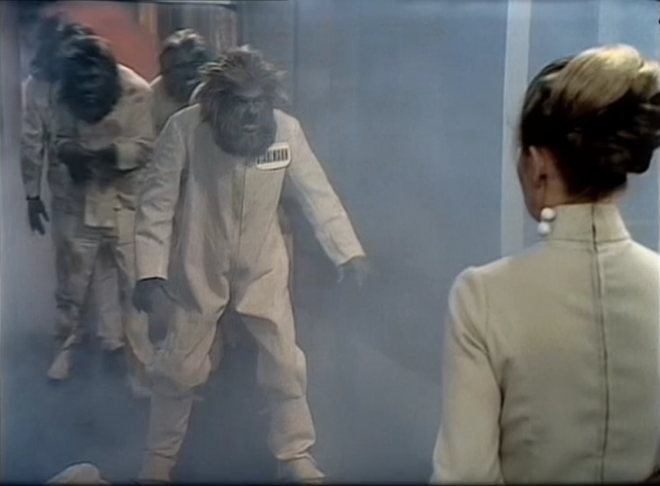 A group of Primords advances on Petra. They have greyish skin and wild hair and beards, and hairy hands too. They're all wearing white overalls. The one at the front has a large nametag reading 'STAHLMAN'