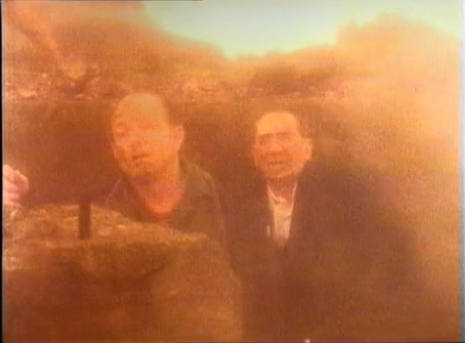 A pair of men sitting amid rubble as the world burns around them with looks of despair on their faces.