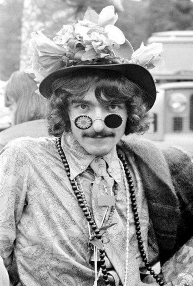 B&W fashion photograph of a white man with a neatly trimmed moustache and a styled mop of curls and a pair of glasses where the lenses have been blacked out, and the right lens has been painted over with a stylized daisy blossom.  He wears multiple long strands of beads as necklaces over a floral/paisley patterned dress shirt with a matching neck-tie