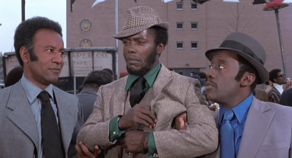 Movie still of three well black men dressed in suits having an engaged conversation on the street