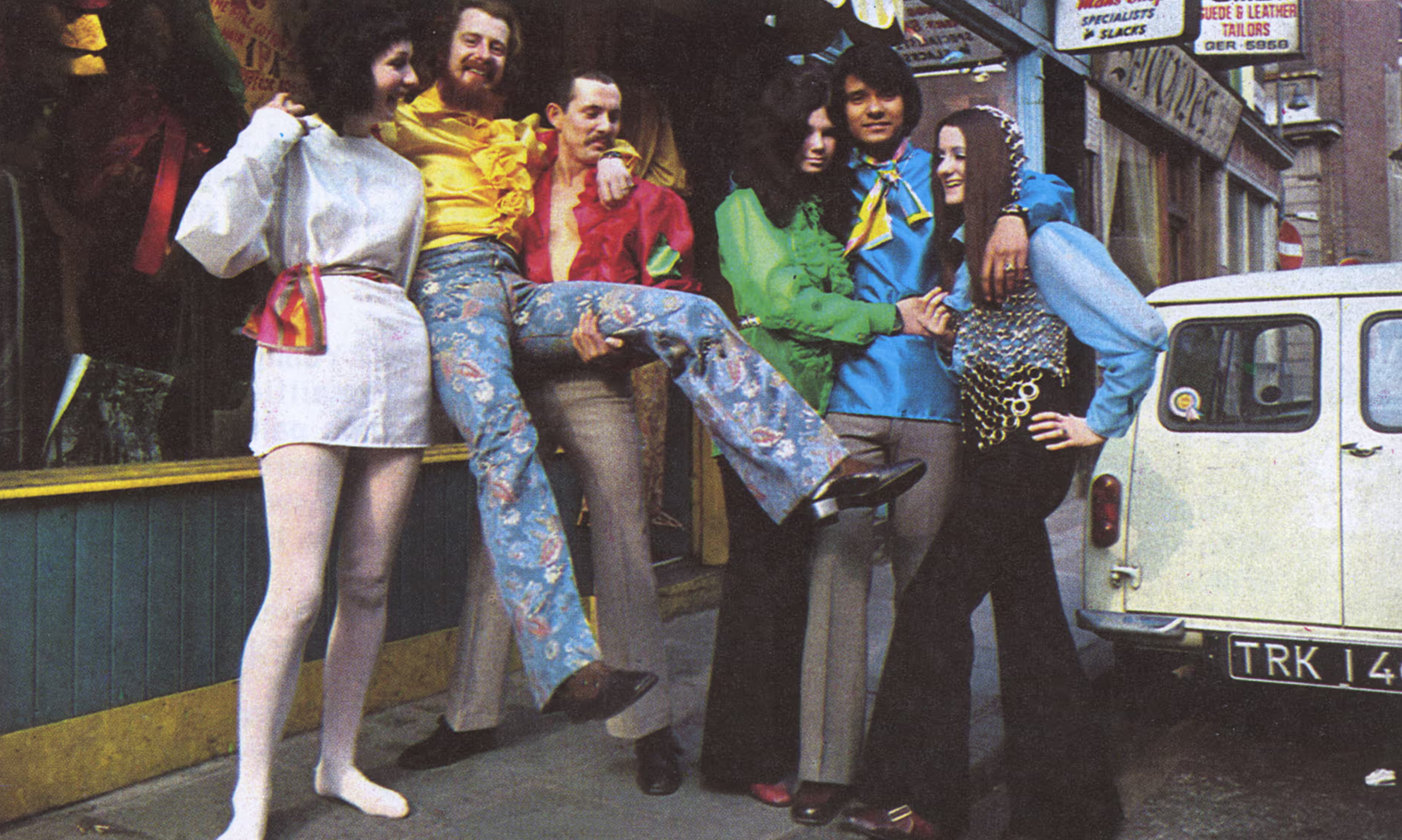 Colour photograph of a brightly dressed sextet of three men and three women ebulliently wearing their home-tailored finery on street.  The two wings of the left trio raise a man with long hair showing off his elaborately painted jeans and well polished footwear