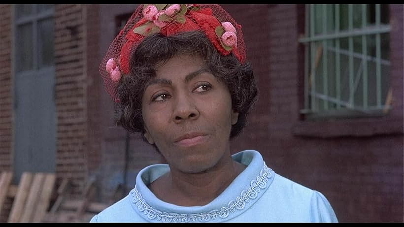Movie still of a middle-aged black woman wearing a hat with lace & flowers looking dubiously on at whatever is taking place