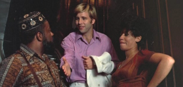 Movie still in which a clean-cut Elgar Enders, looking somewhat awkward but attempting to put on a social face, is caught in the act of introducing himself to a Black man and woman who flank him in the hallway