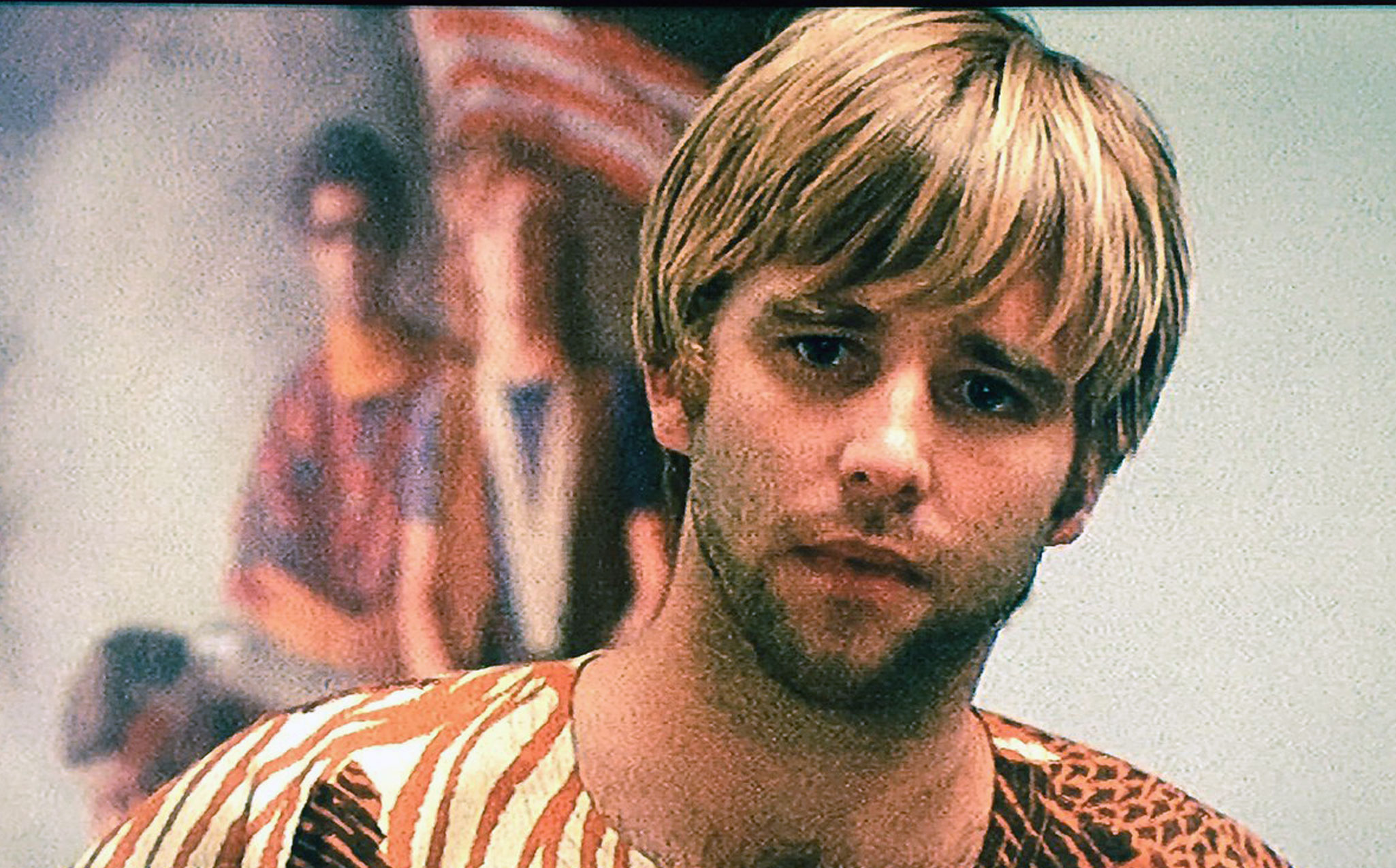 Close-up still of Elgar wearing an African printed top with a concerned and pensive look on his face with what appears to be a group of protesters carrying an American flag in the unfocused background