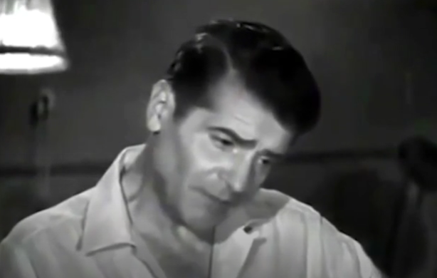 A faintly blurry image of a dark-haired man with a largely unbuttoned shirt, standing next to a lamp.