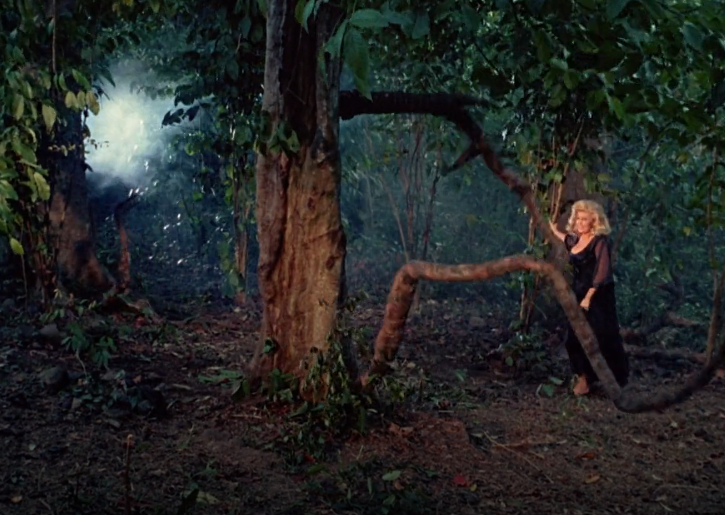 A color image of a woman being lightly menaced by a fake tree root. The wires are clearly visible, as is daylight through the trees.