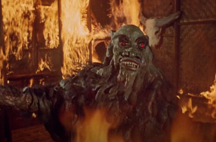 Color image of the monster in a burning hut. The monster is hunched over with the weight of the suit, hands flung wide. Its eyes are red, its skin is rubbery with sculpted tentacles, and its face is still and masklike.