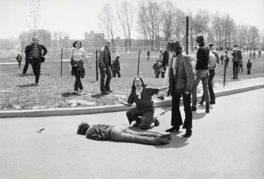 B&W photograph of a dark-haired white teenager kneeling in the road next to a prone body with her arms outstretched and her face raised in shock and grief, as a crowd of concerned people begins to re-congregate