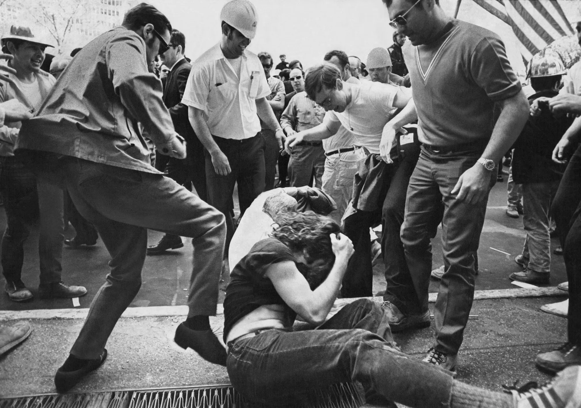 B&W photograph of clean-cut white men in belted trousers wearing sunglasses and hard-hats kicking at a smaller-group of long-haired, jeans-wearing people sitting hunched protectively on the ground