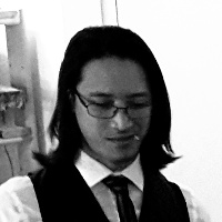 Black and white photograph of a besuited and clean-shaven young Vietnamese man with dark, shoulder-length hair wearing glasses looking at something below the camera and grinning