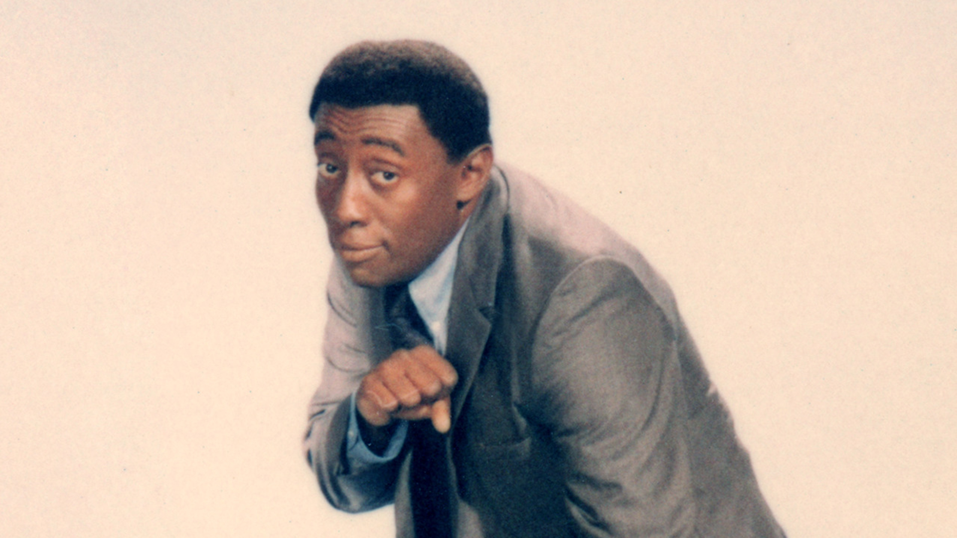 Picture of a young Black man with wearing a suit, ducking slightly and angled to the right, looking at the camera playfully, posed as though preparing to throw a punch with his right hand