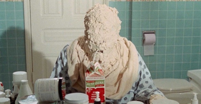 Movie still showing Jeff seated with a drop-cloth draped over his shoulders and thick white paste slathered covering his head to the point of anonymity, sipping from a milk carton with a straw