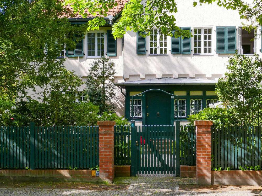 A color photograph of a white plaster two-story building with trees in its front yard. The upper floor has multi-paned windows with dark green shutters. The bottom floor has wooden accents around the windows and door in the same green. A green picket fence with brick pillars on either side of the gate is in the foreground.