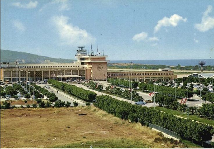 A color photo of Beirut international airport, surrounded by a parking lot with green hedges separating the rows. A brown field is in the foreground. The sky is blue with small wispy clouds.