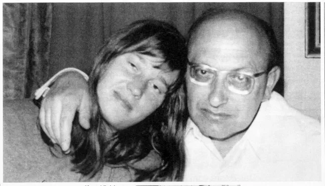 A black and white candid photo of a white woman and a white man standing close to each other and smiling at the camera. The woman's head is leaning on the man's, and he has his arm over her shoulder.