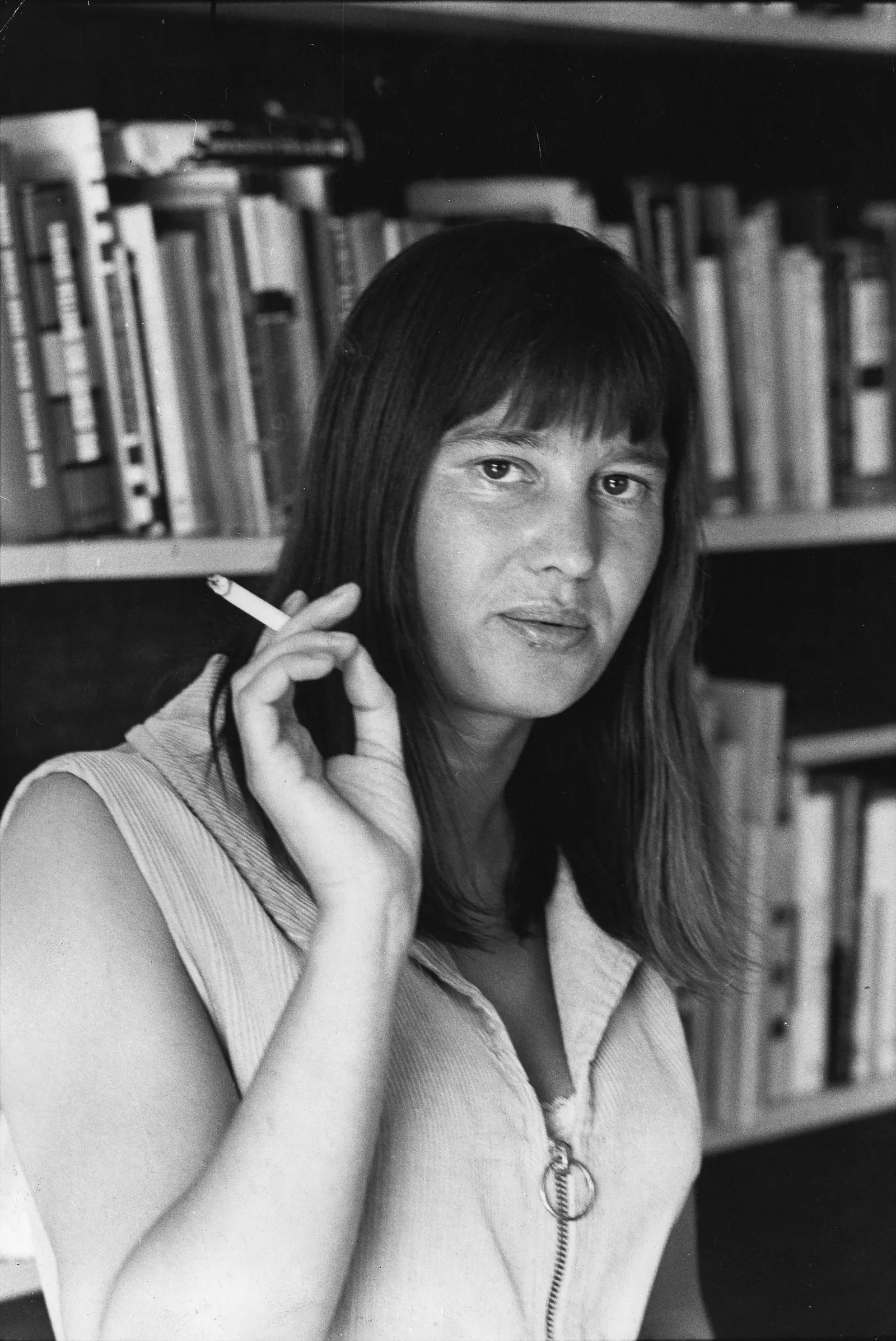 A black and white photo of a white woman with dark shoulder-length hair. She is wearing a white sleeveless zip-up blouse and holding up a cigarette as though she had just taken a drag. She is standing in front of a full bookshelf and looking at the viewer with a neutral expression.