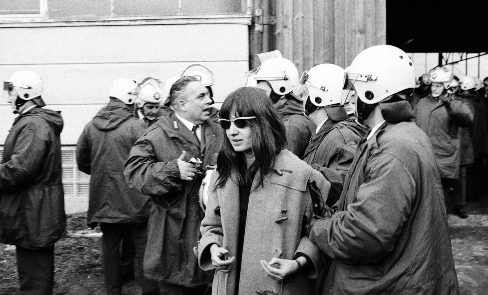 A black and white photo showing Ulrike Meinhof wearing sunglasses and an overcoat, surrounded by police in uniform jackets and helmets. One of the police is holding her arm. A bareheaded man in a suit and tie is speaking to the officers nearest Meinhof.
