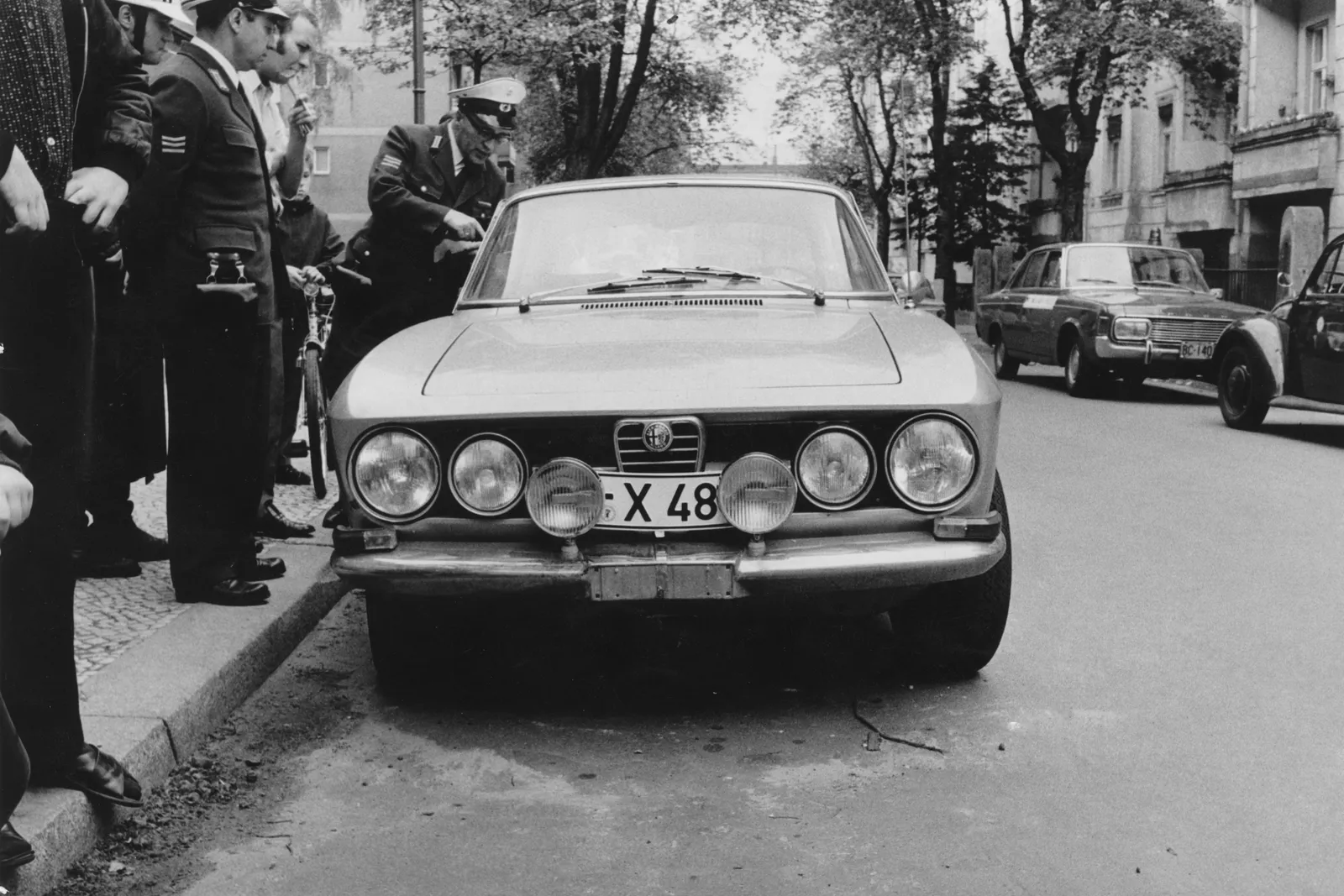 A black and white photo of a city street with parked cars along both sides. The getaway car is at the center of the photo, and to the left a line of police officers stand on the sidewalk looking at it.
