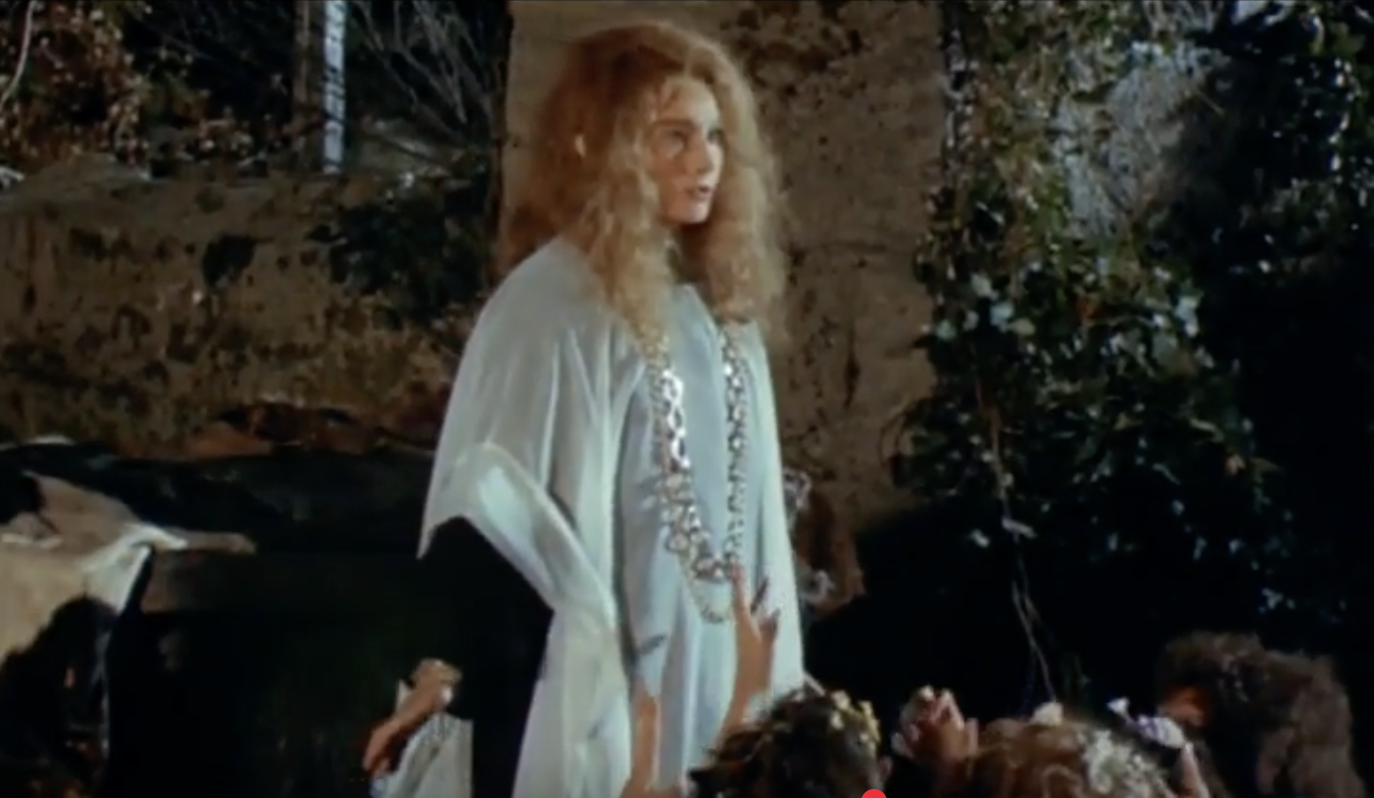 An older blonde woman in a white gown and floral necklace; many hands reach up to touch her.