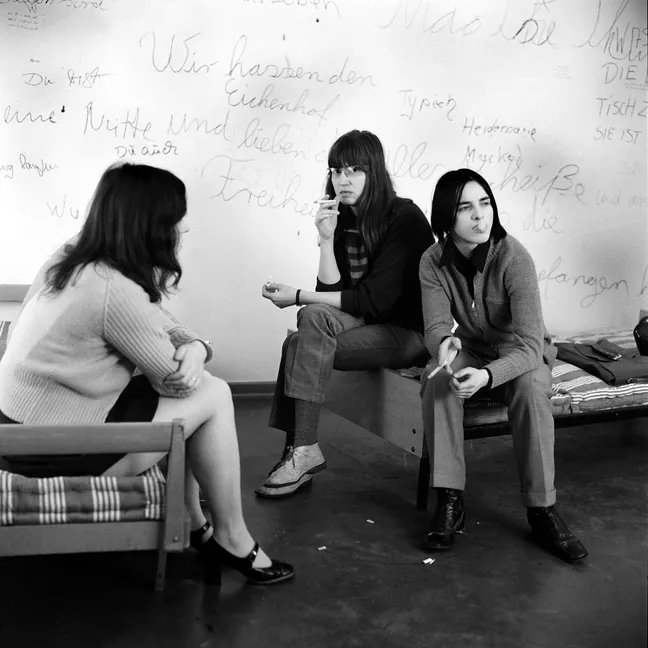 A black and white photo of three women sitting on thin striped mattresses on wire bedframes. Behind them is a wall covered in German graffiti written in youthful handwriting. Meinhof is to the left, facing away from the viewer and toward the other two women. Goergens is in the center, facing Meinhof but looking at the viewer, and holding a cigarette to her lips. The third woman sits next to Goergens facing the viewer but looking away out of the frame. She holds a cigarette in her hand and is exhaling smoke.
